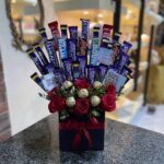A black square box filled with an arrangement of various chocolate bars, golden Ferrero Rocher chocolates, and red roses, tied with a red ribbon