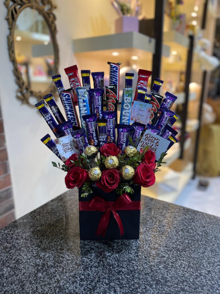 A black square box filled with an arrangement of various chocolate bars, golden Ferrero Rocher chocolates, and red roses, tied with a red ribbon