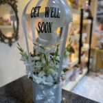 A clear balloon with the words "GET WELL SOON" printed on it, floating above an arrangement of white flowers in a light grey container adorned with a silver bow.