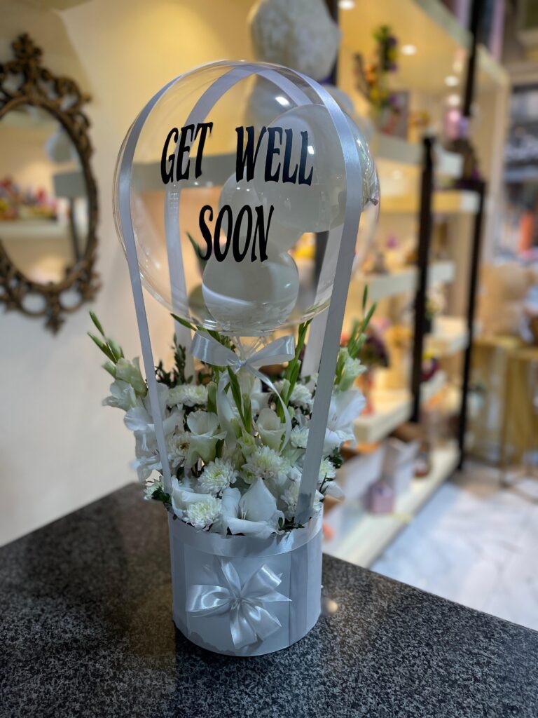 A clear balloon with the words "GET WELL SOON" printed on it, floating above an arrangement of white flowers in a light grey container adorned with a silver bow.