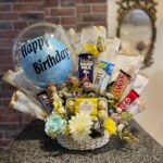 A white wicker basket filled with a clear "Happy Birthday" balloon, an assortment of chocolate bars, and small flowers