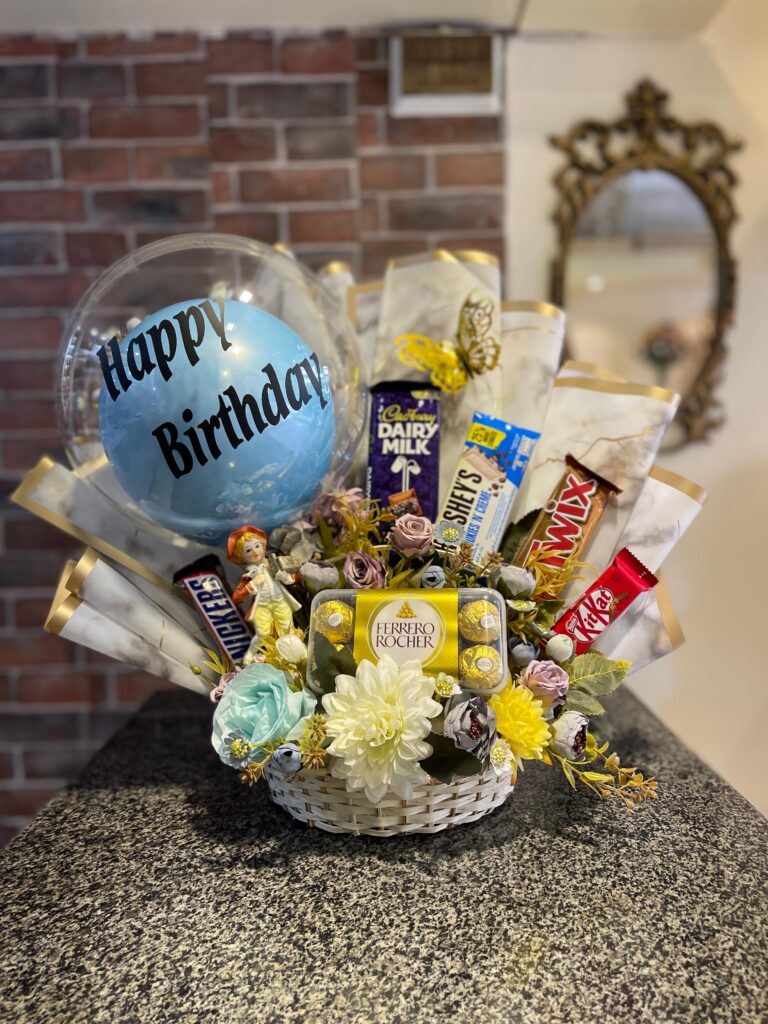 A white wicker basket filled with a clear "Happy Birthday" balloon, an assortment of chocolate bars, and small flowers