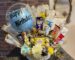 A white wicker basket filled with a clear "Happy Birthday" balloon, an assortment of chocolate bars, and small flowers