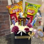 A black gift box with a gold ribbon and bow, containing a bouquet of white and purple flowers and an assortment of snacks and chocolate bars, including two bags of Lay's potato chips.