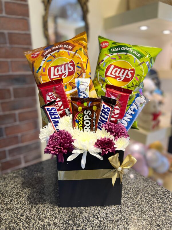 A black gift box with a gold ribbon and bow, containing a bouquet of white and purple flowers and an assortment of snacks and chocolate bars, including two bags of Lay's potato chips.