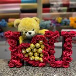 A gift box spelling "I ❤️ U" with red roses, a heart filled with Ferrero Rocher chocolates, and a yellow teddy bear. The box is on a speckled countertop