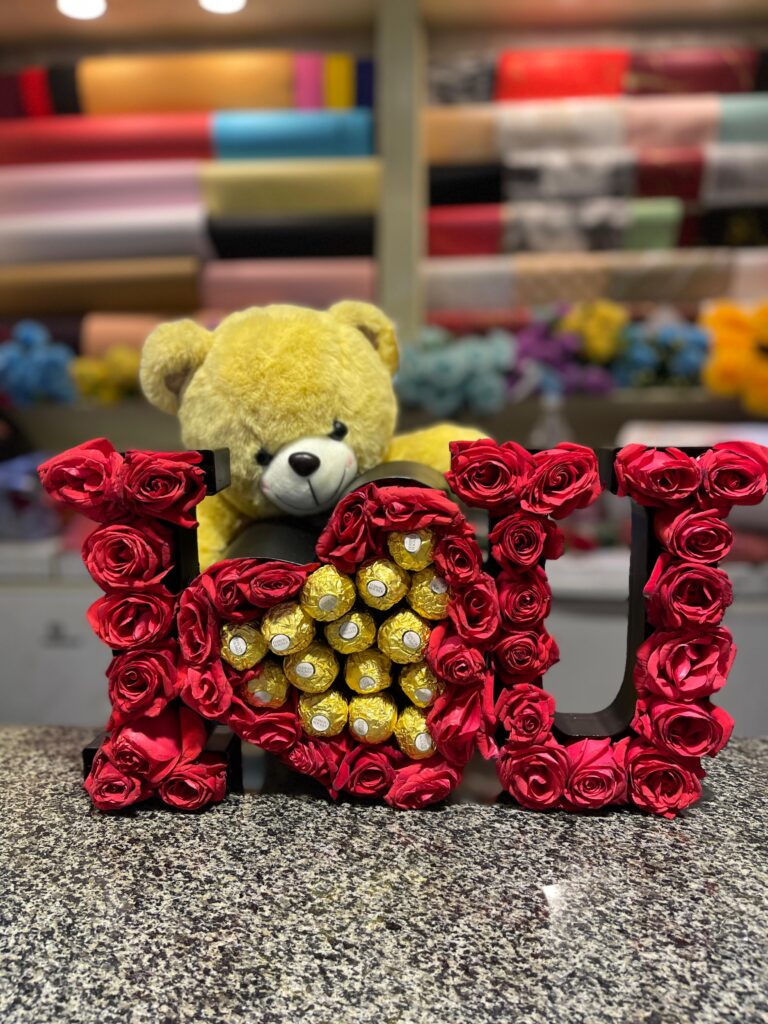 A gift box spelling "I ❤️ U" with red roses, a heart filled with Ferrero Rocher chocolates, and a yellow teddy bear. The box is on a speckled countertop